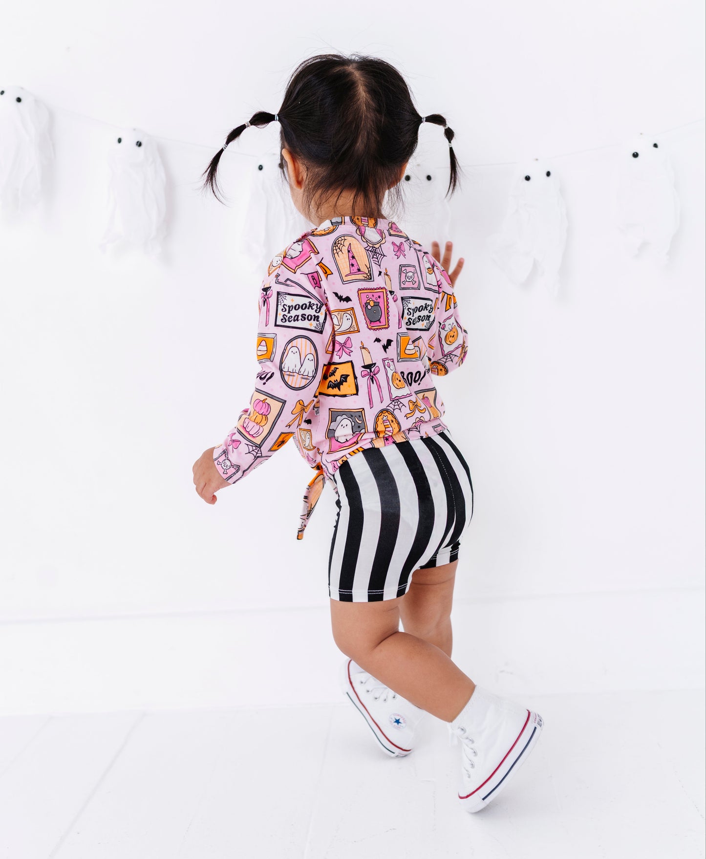 A toddler girl is shown from behind wearing a pink long-sleeve top with a colorful patchwork graphic print and black-and-white striped shorts, standing against a white wall with ghost decorations.