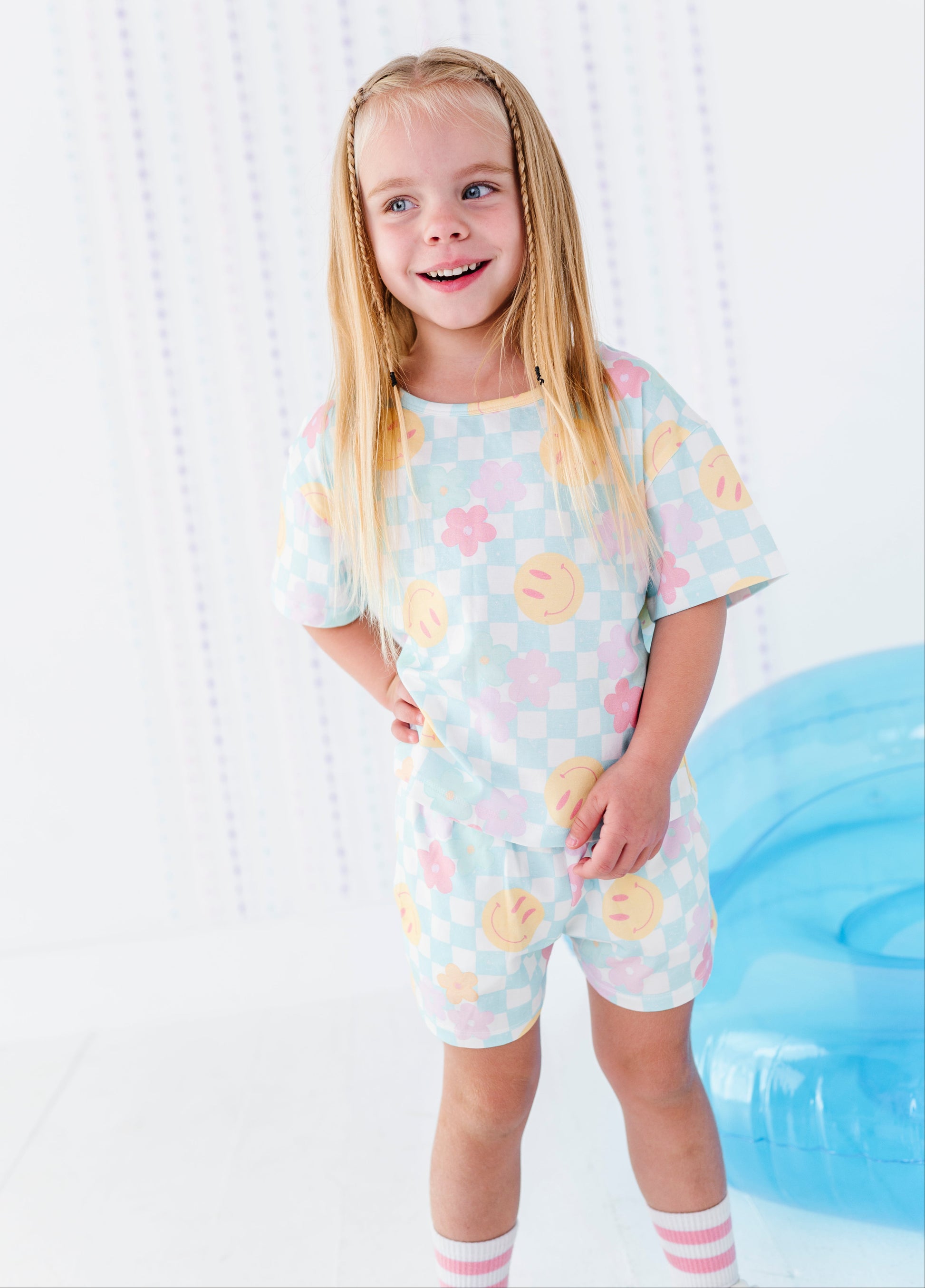 A young girl with long blonde hair stands smiling, wearing a pastel checkered short-sleeve pajama set with a smiley face pattern. The set features a light blue and white checkerboard base with pink, yellow, and peach smiley faces.