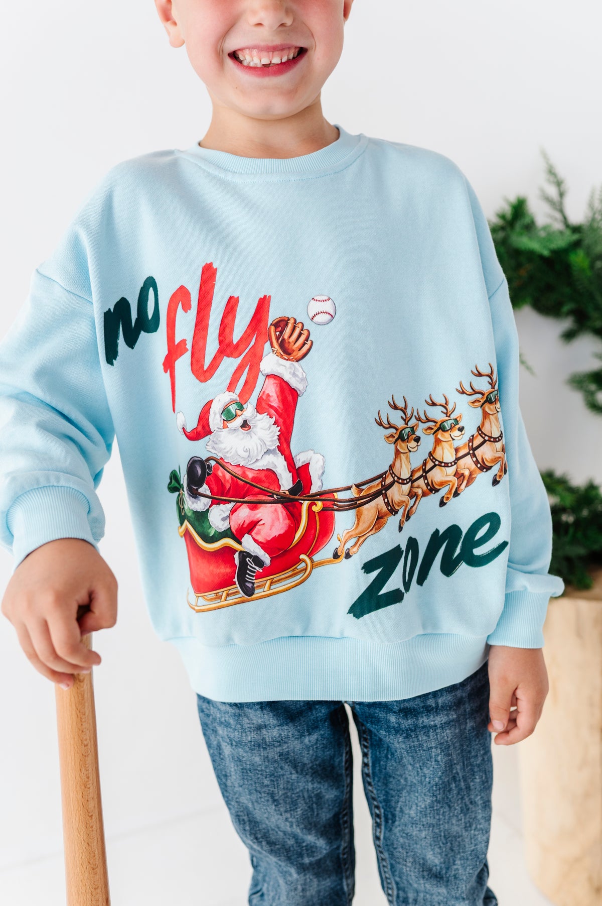 A close-up detail shot of the blue "no fly zone" crewneck. The young boy wearing the crewneck is also holding a baseball bat and smiling 