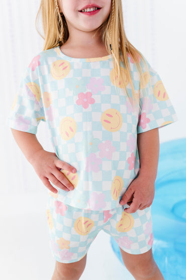 A young girl wearing a matching two-piece pajama set with a pastel checkered pattern and smiley face and flower prints, standing in a brightly lit room.
