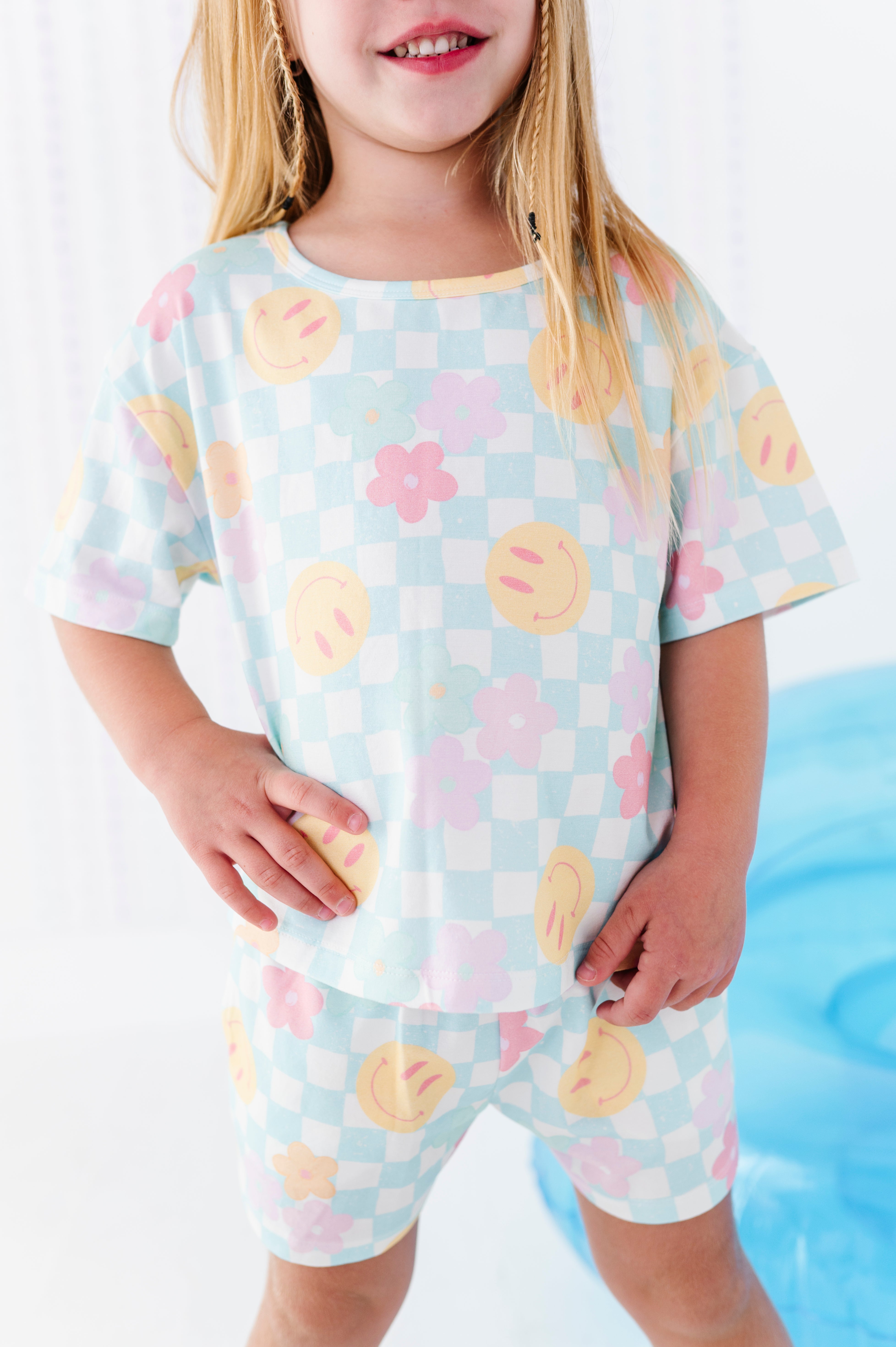 A young girl wearing a matching two-piece pajama set with a pastel checkered pattern and smiley face and flower prints, standing in a brightly lit room.
