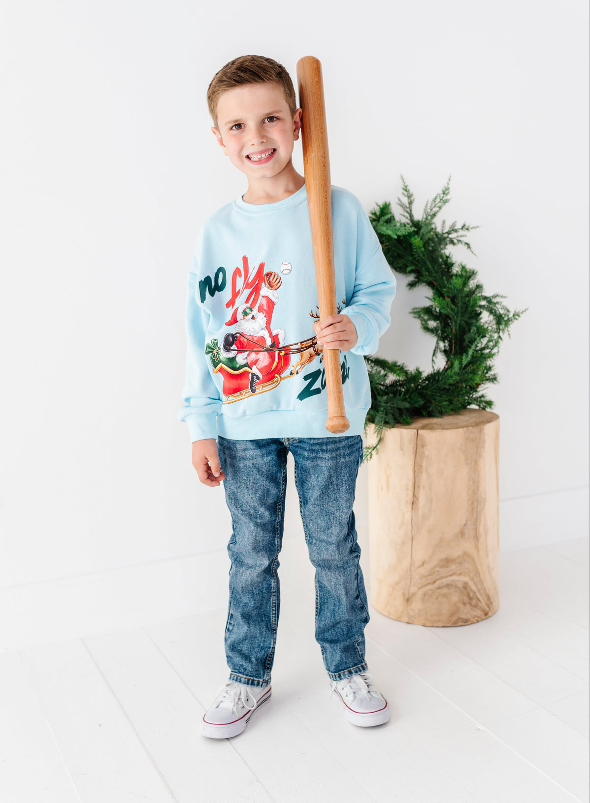 A young boy stands in a bright studio wearing a light blue crewneck sweatshirt with a colorful graphic print, blue jeans, and white sneakers, holding a wooden baseball bat.