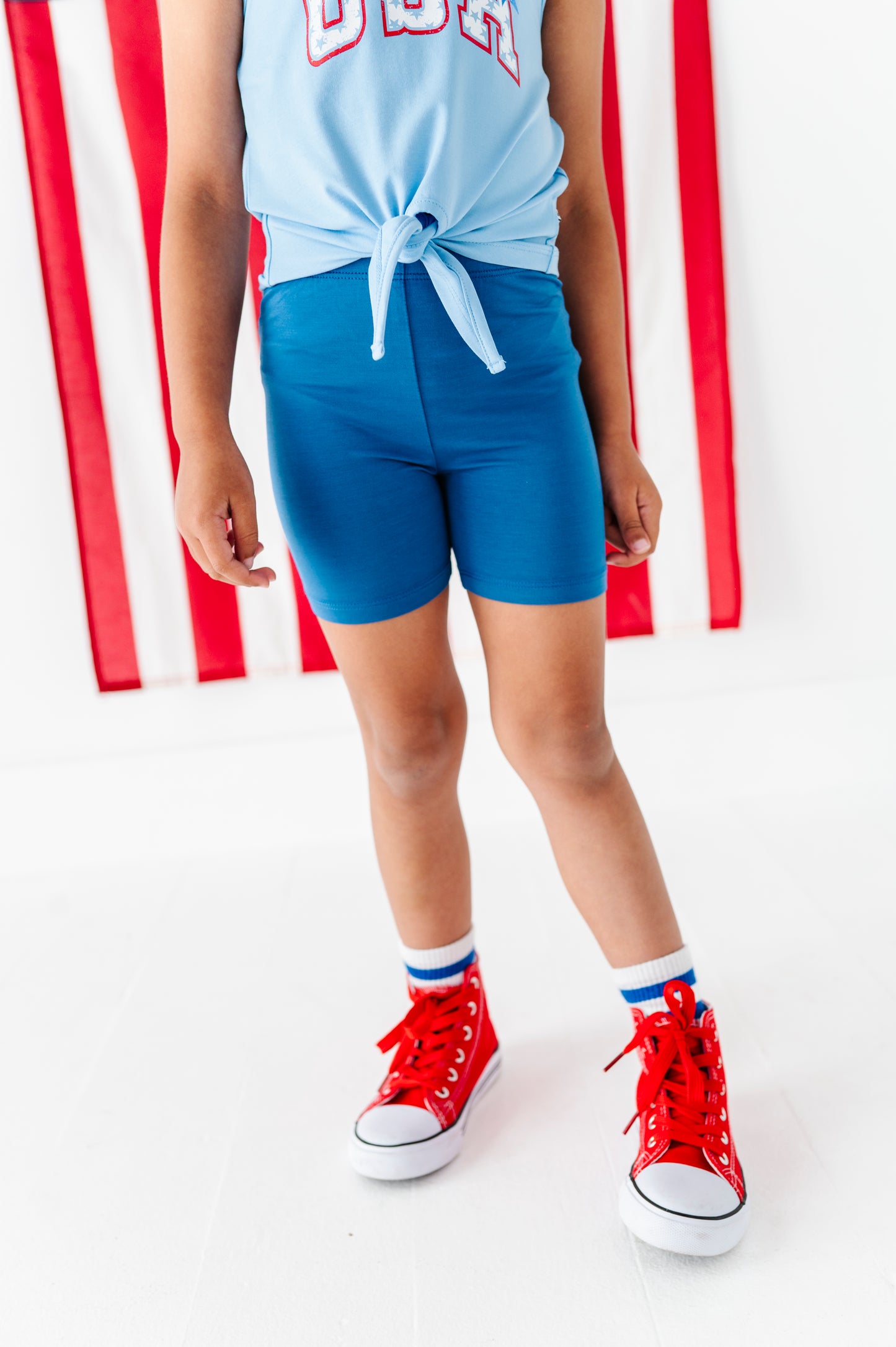 Young girl wearing a light blue sleeveless top and shorts with red high-top sneakers in front of a red and white striped background.