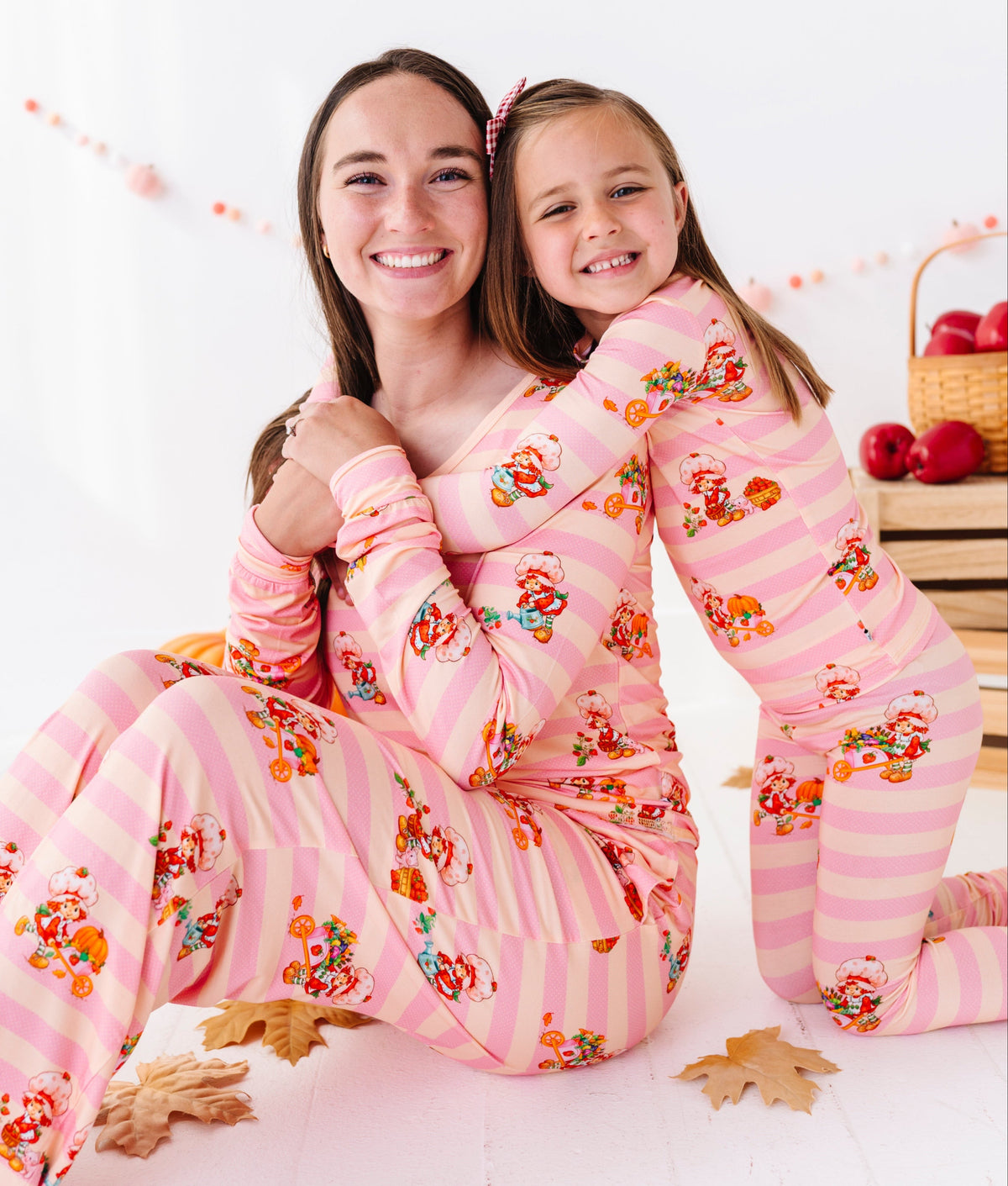 A woman and a young girl hugging while wearing matching pink pajamas with orange and yellow cartoon characters and small floral details. The photo is taken in a bright, white room with decorative autumn leaves and a basket of red apples in the background.