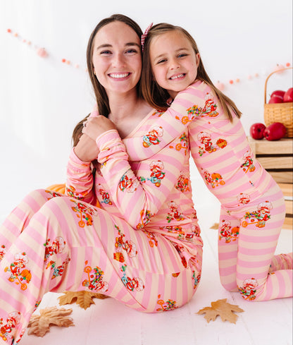 A woman and a young girl hugging while wearing matching pink pajamas with orange and yellow cartoon characters and small floral details. The photo is taken in a bright, white room with decorative autumn leaves and a basket of red apples in the background.