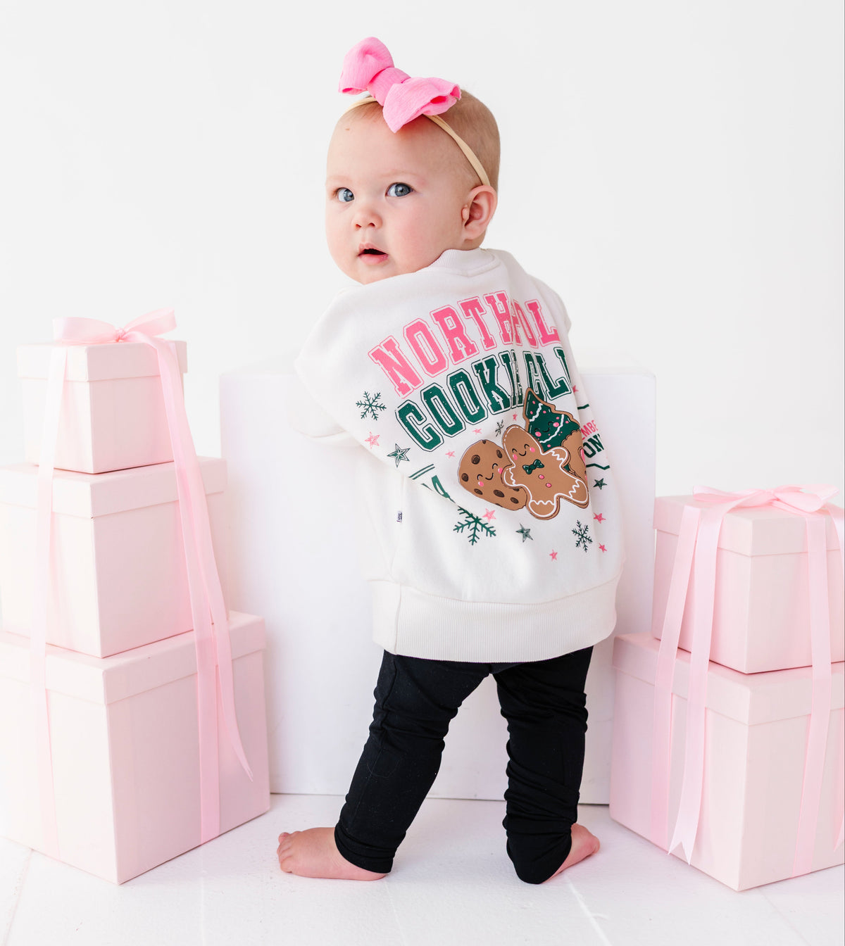 A baby standing on a white floor wearing a white graphic crewneck with 'NORTH POLE COOKIE CLUB' and a gingerbread cookie design, black leggings, and a pink bow headband. The baby is posed between stacks of pink gift boxes with pink ribbons.