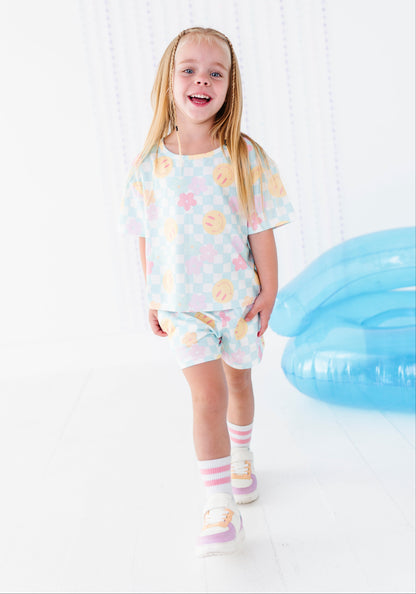 A young girl with long blonde hair is smiling and walking forward in a matching pastel checkered t-shirt and shorts set. The outfit features a light blue, pink, and yellow pattern.
