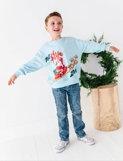 A young boy wearing a light blue bamboo "no fly zone" crewneck and jeans. He stands in front of a simple green wreath and a tree stump 