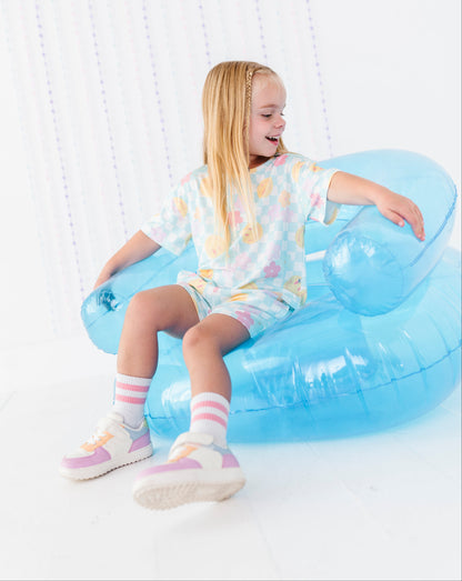 A young girl with long blonde hair sits smiling on a light blue inflatable pool float chair, wearing a pastel multicolor tie-dye t-shirt and shorts with pink and white striped socks and pastel sneakers.
