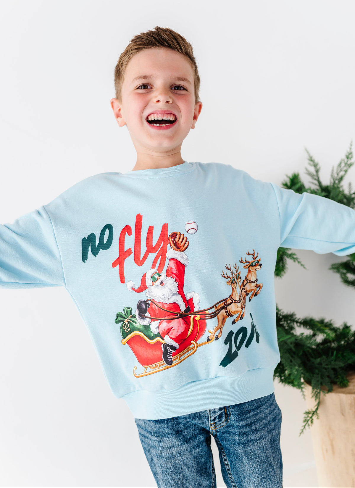 A young boy smiling and laughing while wearing a light blue crewneck sweatshirt with a festive graphic of Santa Claus in a sleigh, the text 'no fly zone'