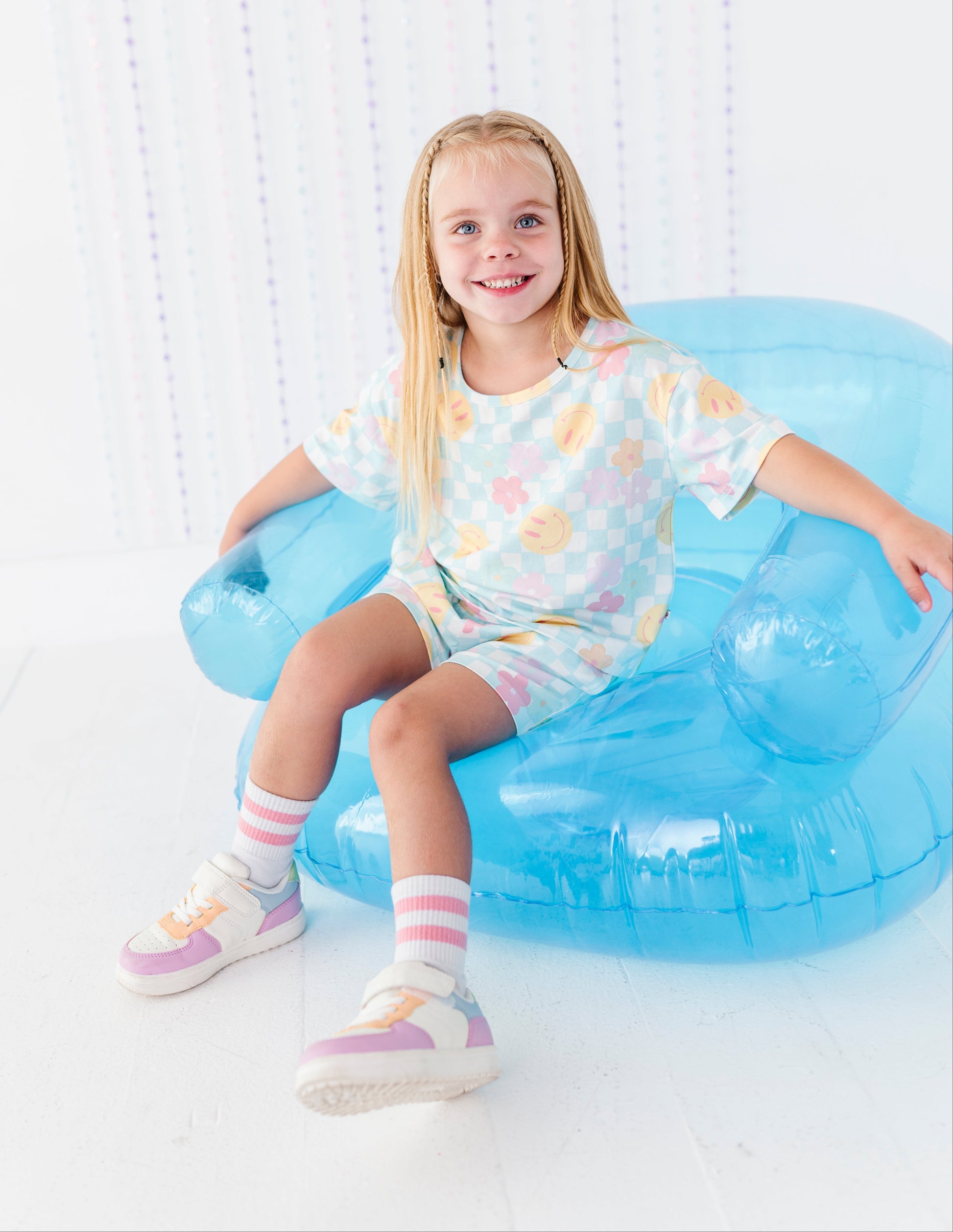 A young girl with blonde hair sits smiling on a translucent light blue inflatable chair. She is wearing a pastel multicolor patterned t-shirt and matching shorts, pink and white striped socks, and pastel sneakers.