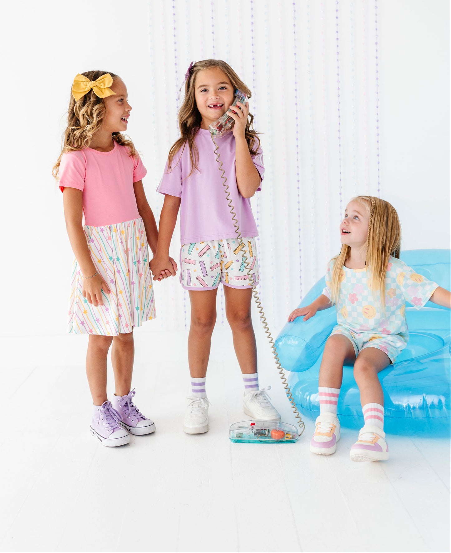 Three young girls in pastel-colored outfits play with a toy telephone in a bright, white room. One girl on the left wears a pink shirt and a light pink patterned skirt, holding hands with the middle girl who wears a lavender shirt and matching pastel shorts.