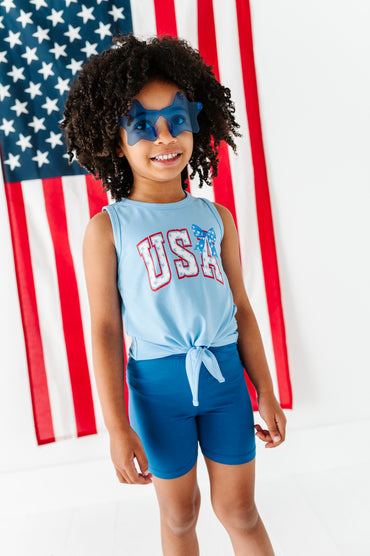 Child wearing a blue 'USA' bamoo tank top and shorts with a superhero mask in front of an American flag.