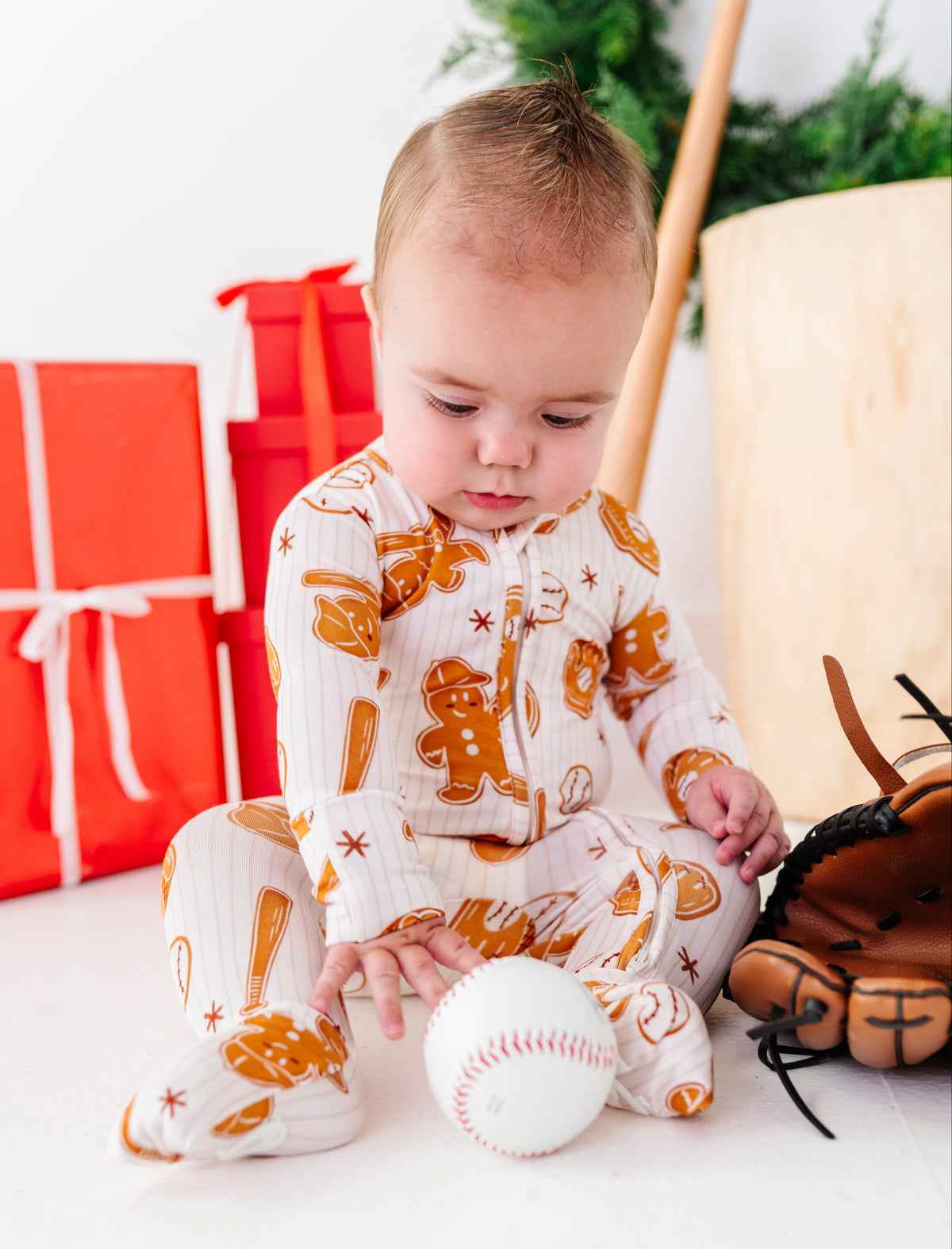 A baby sitting on a white floor wearing a white and orange gingerbread-themed footed pajama, touching a baseball. A baseball glove, wooden bat, and red wrapped gifts are visible in the background.