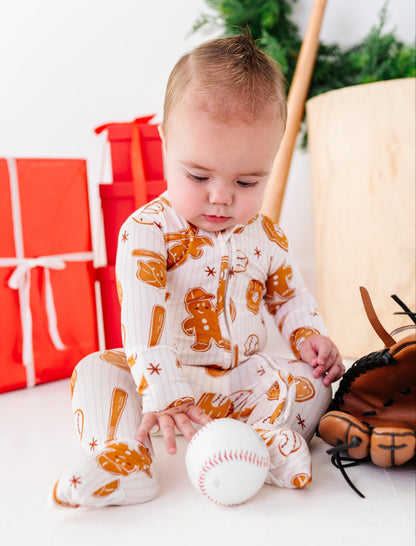 A baby sitting on a white floor wearing a white and orange gingerbread-themed footed pajama, touching a baseball. A baseball glove, wooden bat, and red wrapped gifts are visible in the background.