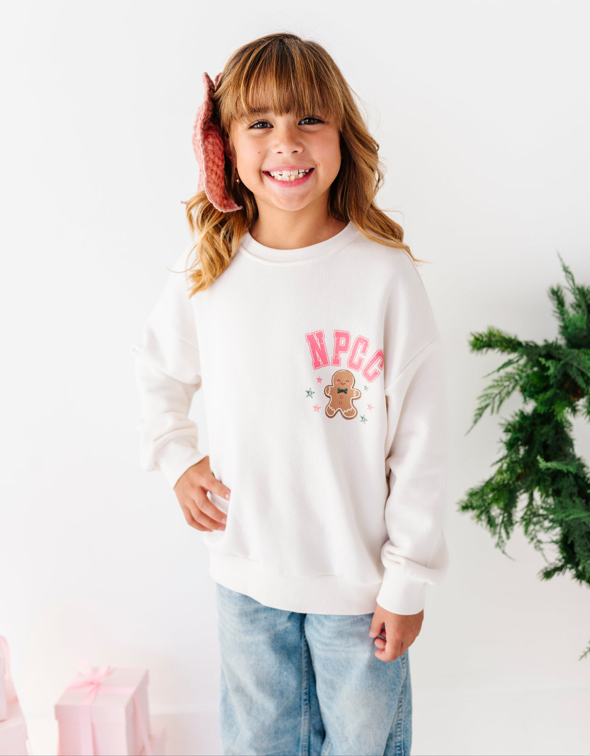 A girl standing on a white floor wearing a white graphic crewneck with 'NPCC' and a gingerbread cookie design, denim jeans, and a pink bow. 