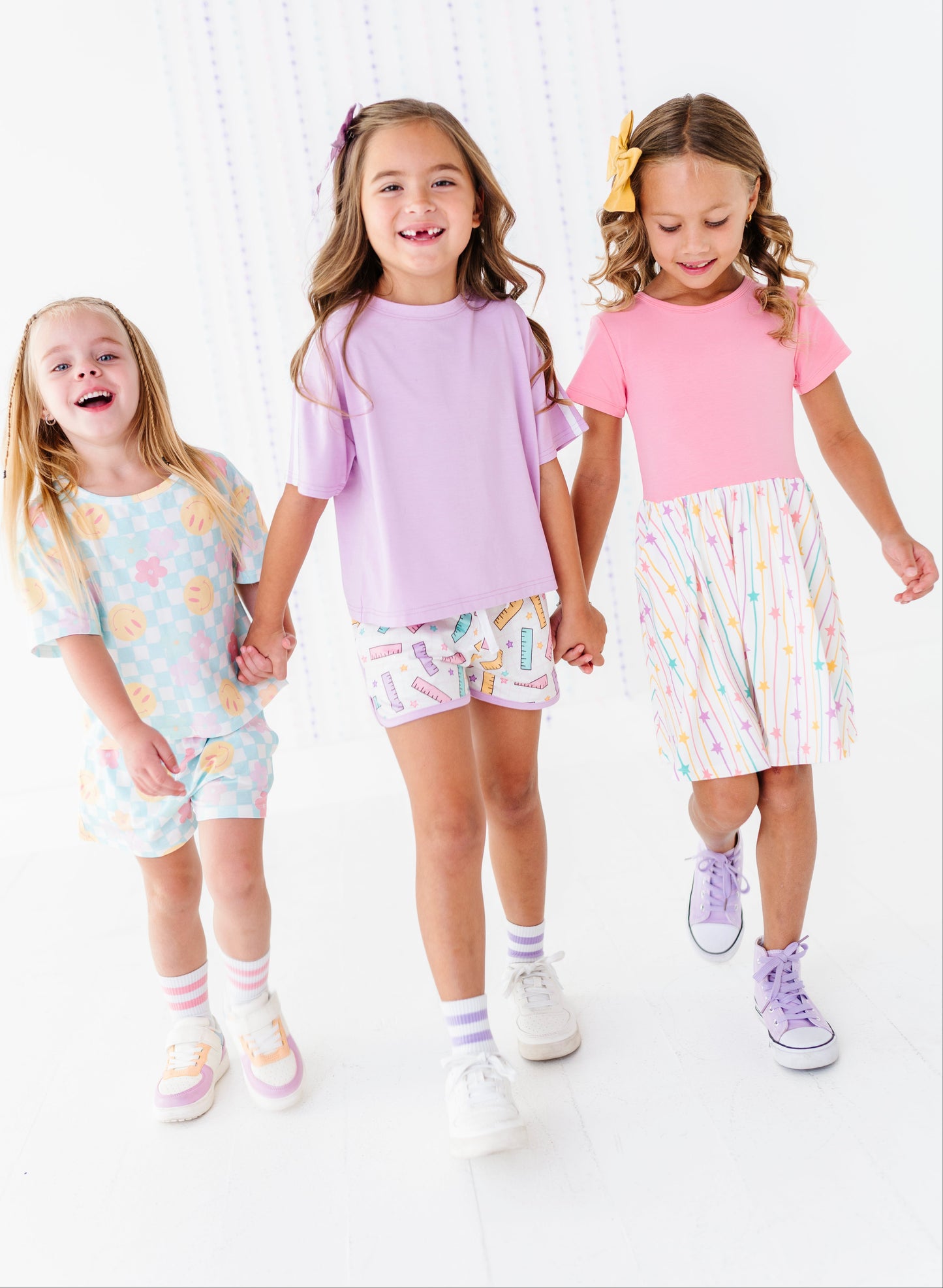Three young girls walking hand-in-hand in a bright, white studio setting, wearing pastel-colored matching outfits. The girl on the left wears a light blue patterned t-shirt and shorts, the middle girl wears a lavender t-shirt and multicolor patterned shorts, and the girl on the right wears a pink t-shirt and a white skirt with pastel vertical stripes.