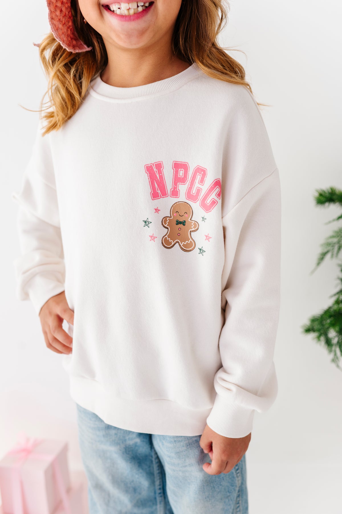An up-close photo of a young girl wearing a bamboo crewneck. The crewneck says "NPCC" above a gingerbread cookie. 