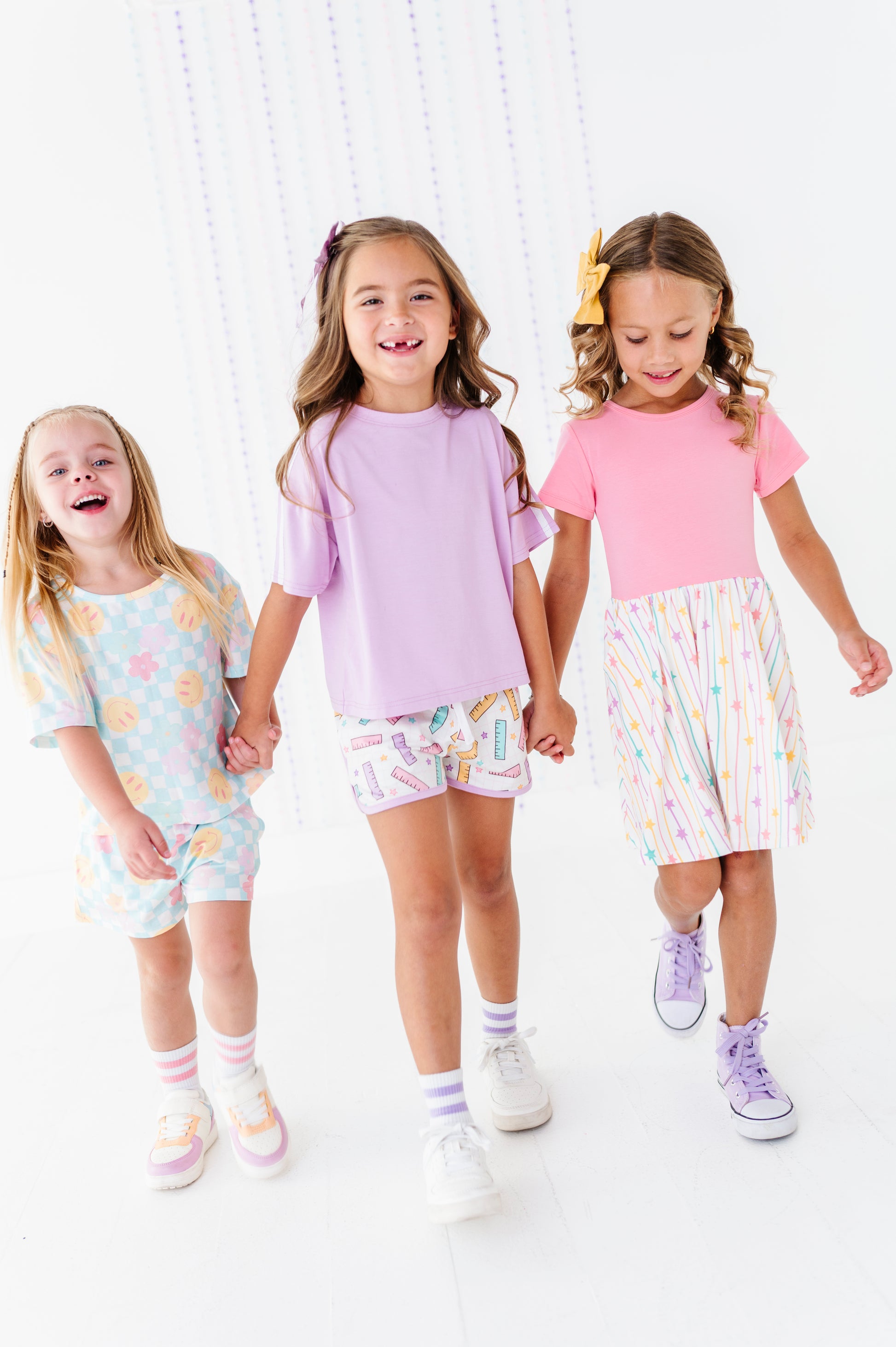 Three young girls walking hand-in-hand in a bright, white studio setting, wearing pastel-colored matching outfits. The girl on the left wears a light blue patterned t-shirt and shorts, the middle girl wears a lavender t-shirt and multicolor patterned shorts, and the girl on the right wears a pink t-shirt and a white skirt with pastel vertical stripes.