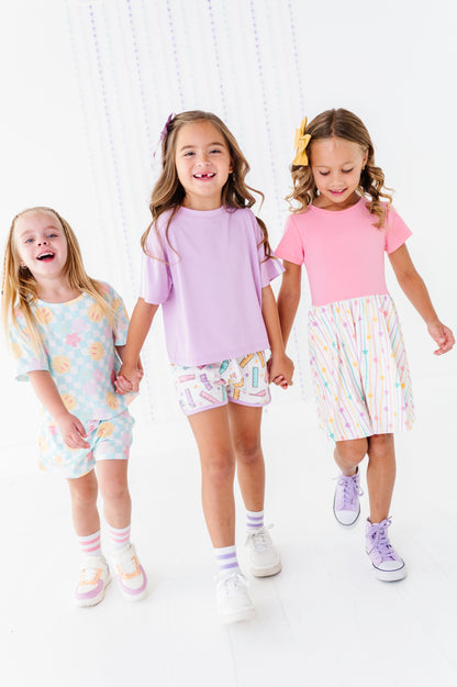 Three young girls walking hand-in-hand in a bright, white studio setting, wearing pastel-colored matching outfits. The girl on the left wears a light blue patterned t-shirt and shorts, the middle girl wears a lavender t-shirt and multicolor patterned shorts, and the girl on the right wears a pink t-shirt and a white skirt with pastel vertical stripes.