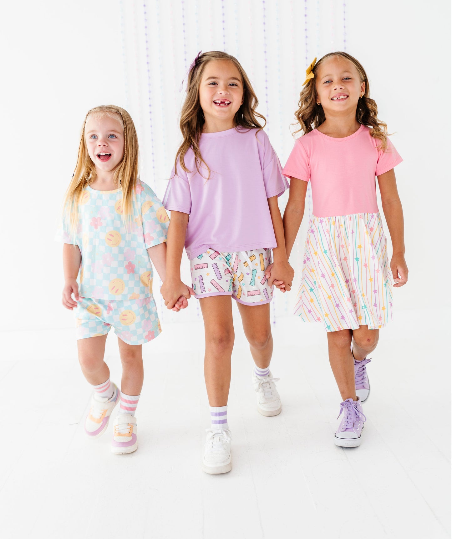 Three young girls walking hand-in-hand in a bright, white studio setting, wearing pastel-colored matching clothing sets. The girl on the left wears a light blue floral romper, the middle girl wears a lavender t-shirt with pastel patterned shorts, and the girl on the right wears a pink t-shirt with a pastel rainbow skirt.
