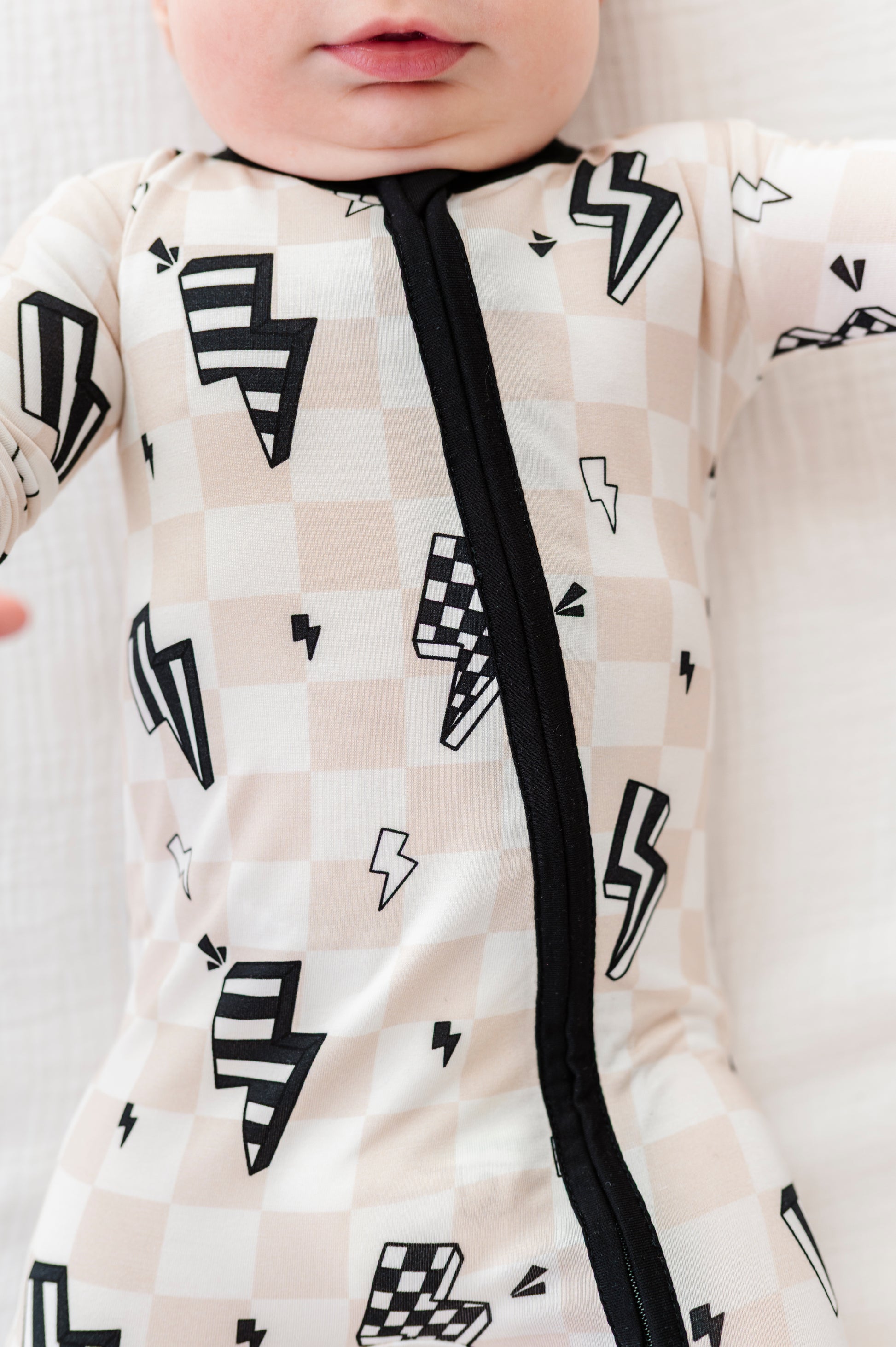 A close-up of a baby wearing a long-sleeve sleep sack with a beige and white checkerboard pattern, black lightning bolt prints, and a black front zipper.