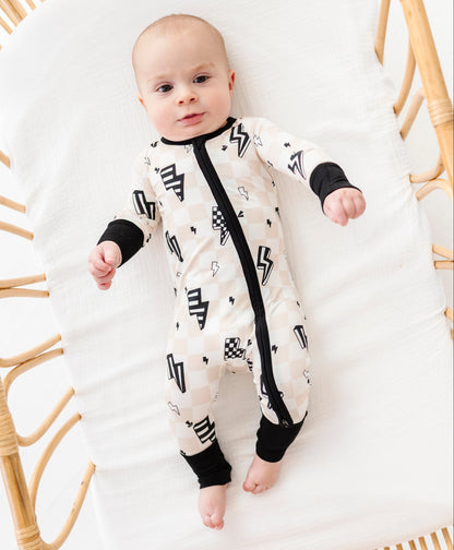 A baby lying on a white cushion inside a rattan bassinet, wearing a cream-colored footless sleeper with black geometric patterns and black trim.