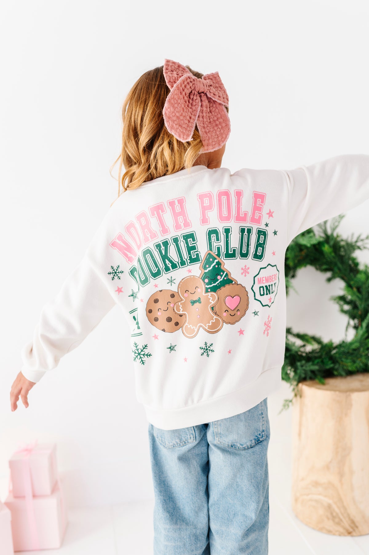 A girl standing on a white floor wearing a white graphic crewneck with 'NORTH POLE COOKIE CLUB' and a gingerbread cookie design, denim jeans, and a pink bow. 