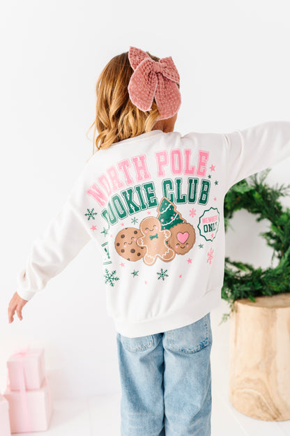 A girl standing on a white floor wearing a white graphic crewneck with 'NORTH POLE COOKIE CLUB' and a gingerbread cookie design, denim jeans, and a pink bow. 