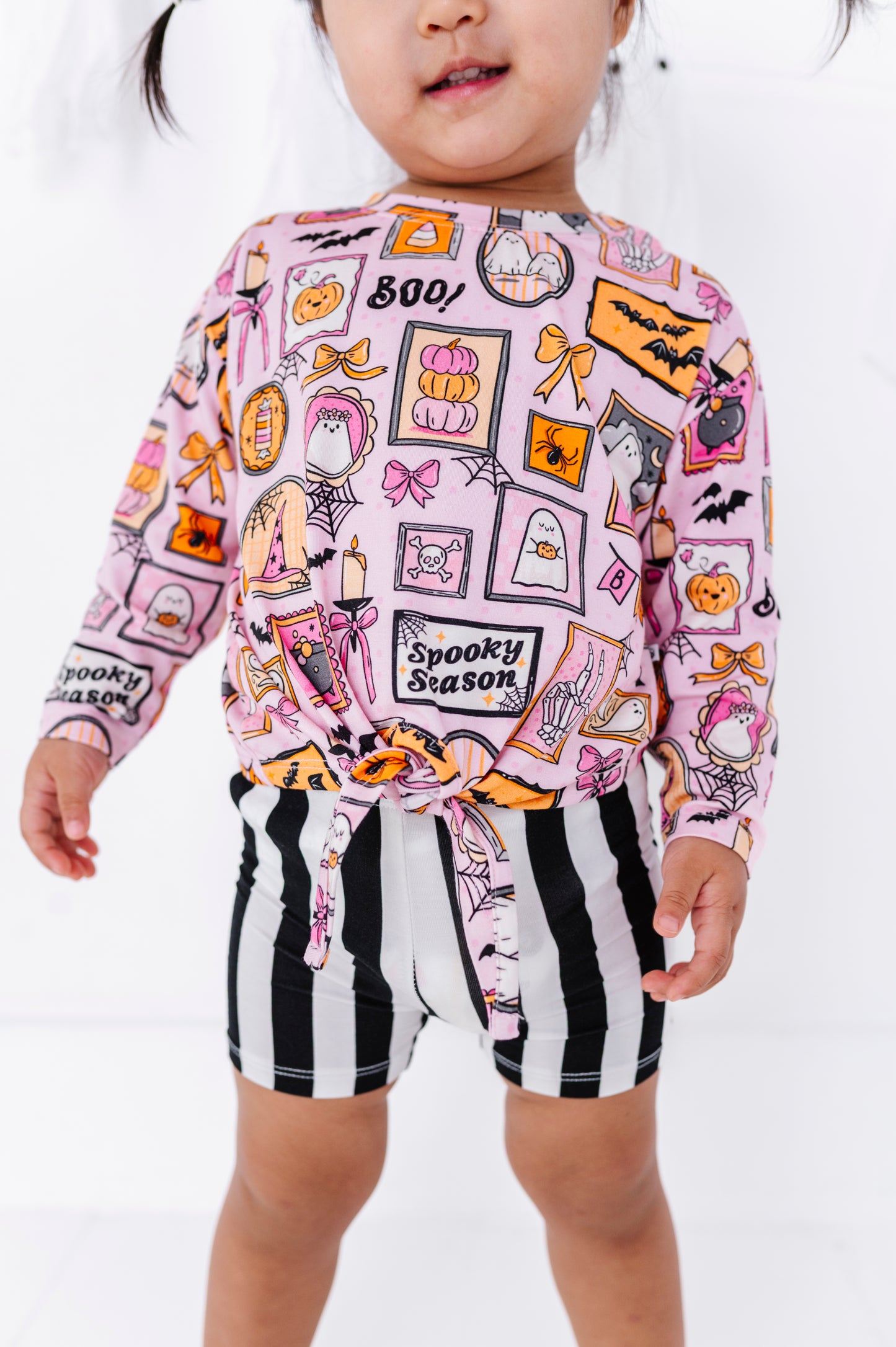 A toddler wearing a pink long-sleeve top with a colorful 'Spooky Season' Halloween print and black-and-white vertically striped shorts, shown against a plain white studio background.