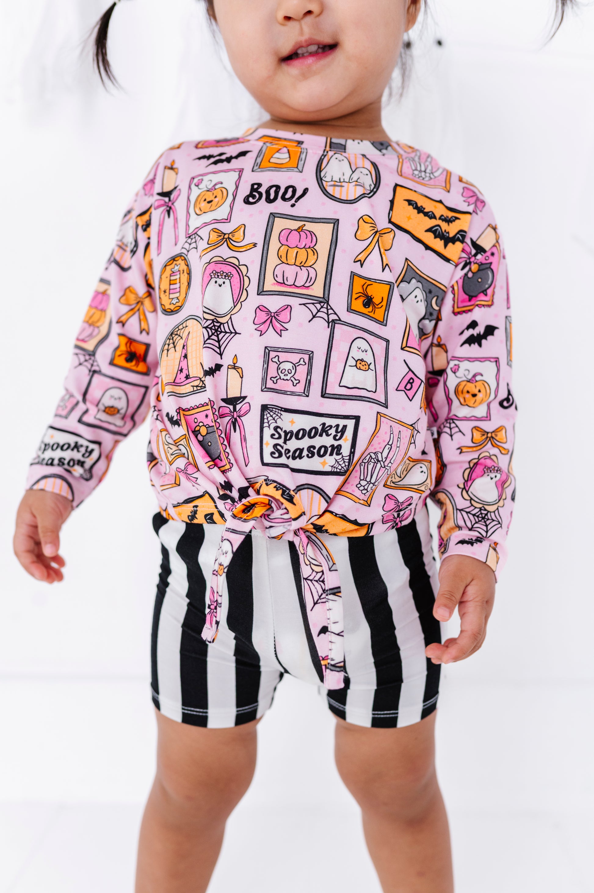 A toddler wearing a pink long-sleeve top with a colorful 'Spooky Season' Halloween print and black-and-white vertically striped shorts, shown against a plain white studio background.