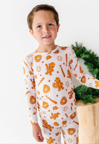 A young boy wearing a white long-sleeve pajama set with an orange baseball-themed print, standing against a white background with a green wreath and wooden box visible behind him.