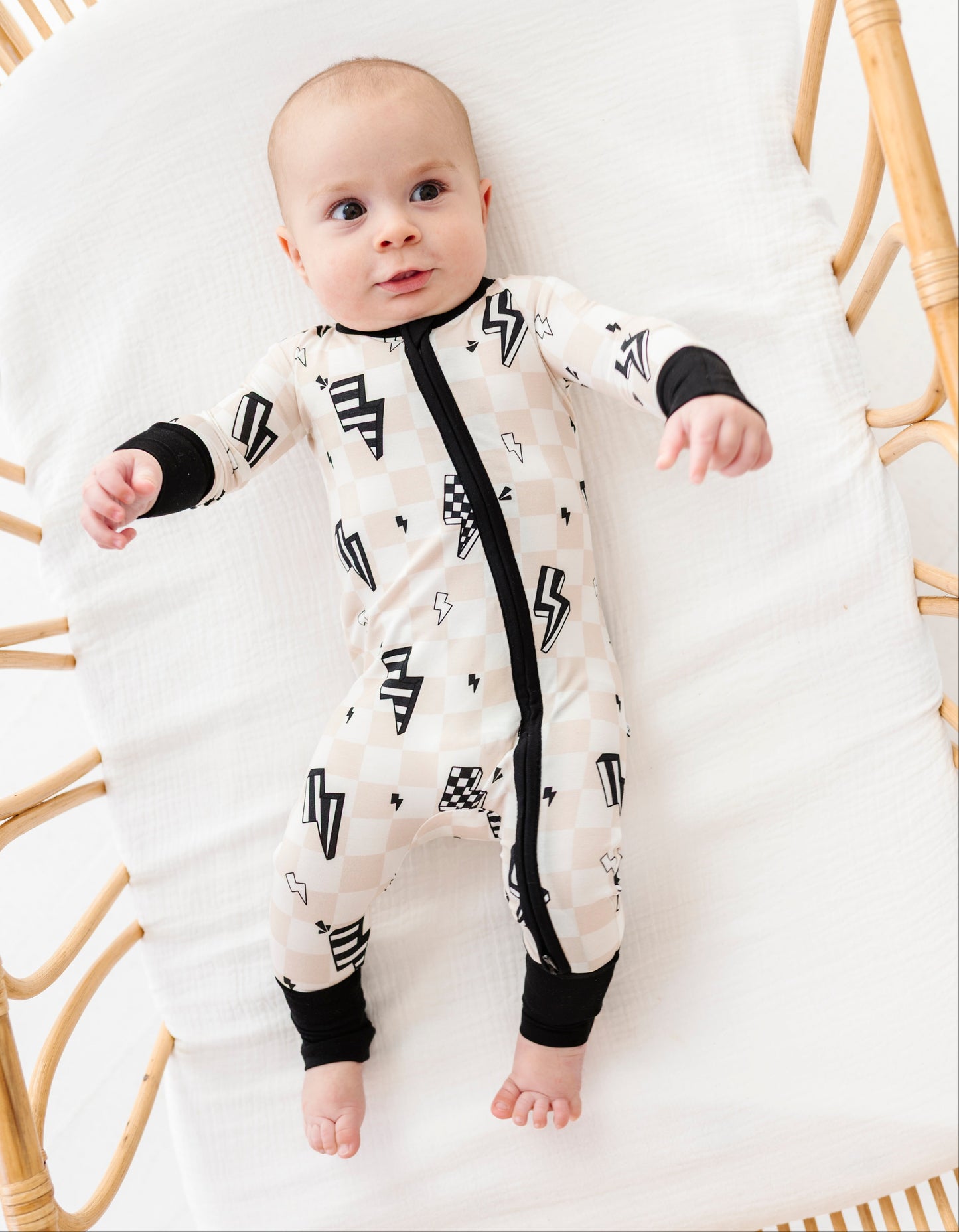 A baby lying on a white muslin bassinet wearing a cream-colored one-piece sleeper with a black zipper, black cuffs, and black geometric patterns.