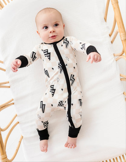 A baby lying on a white muslin bassinet wearing a cream-colored one-piece sleeper with a black zipper, black cuffs, and black geometric patterns.