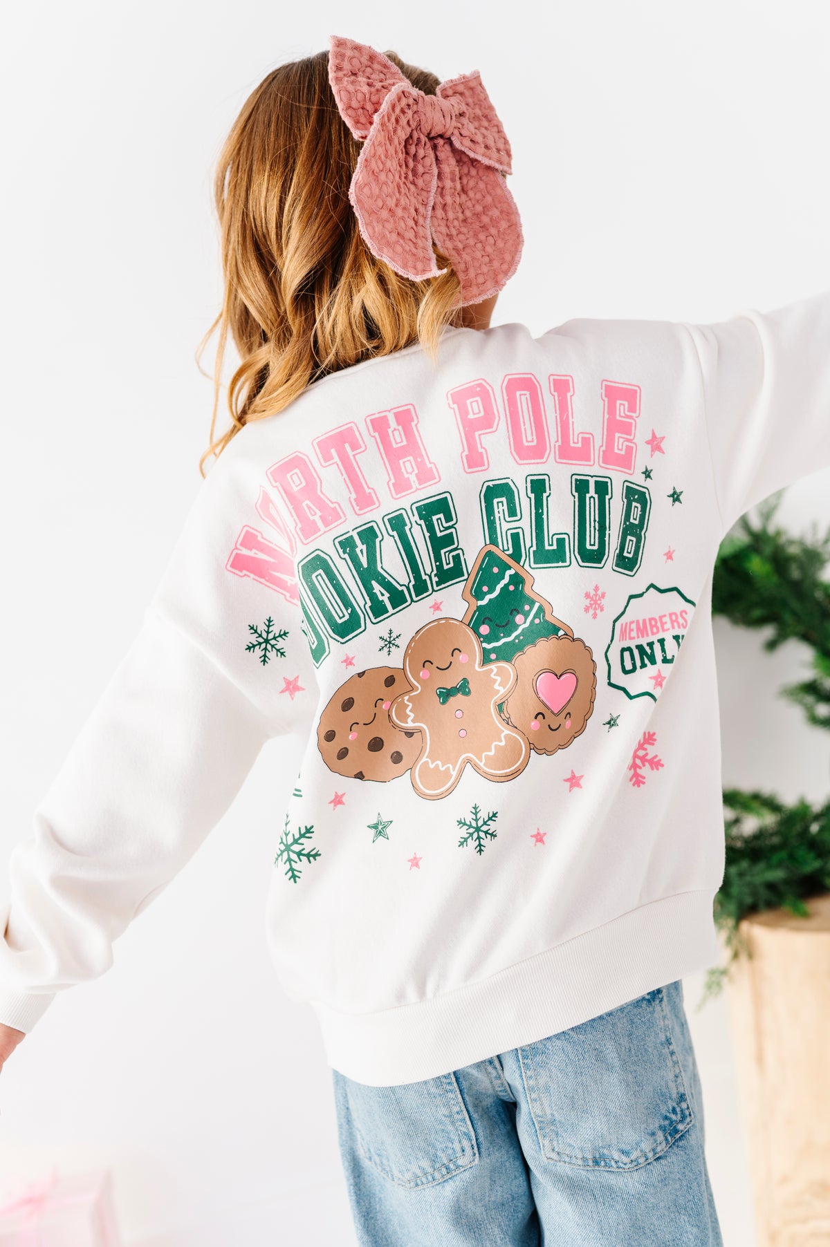A girl standing on a white floor wearing a white graphic crewneck with 'NORTH POLE COOKIE CLUB' and a gingerbread cookie design, denim jeans, and a pink bow. 