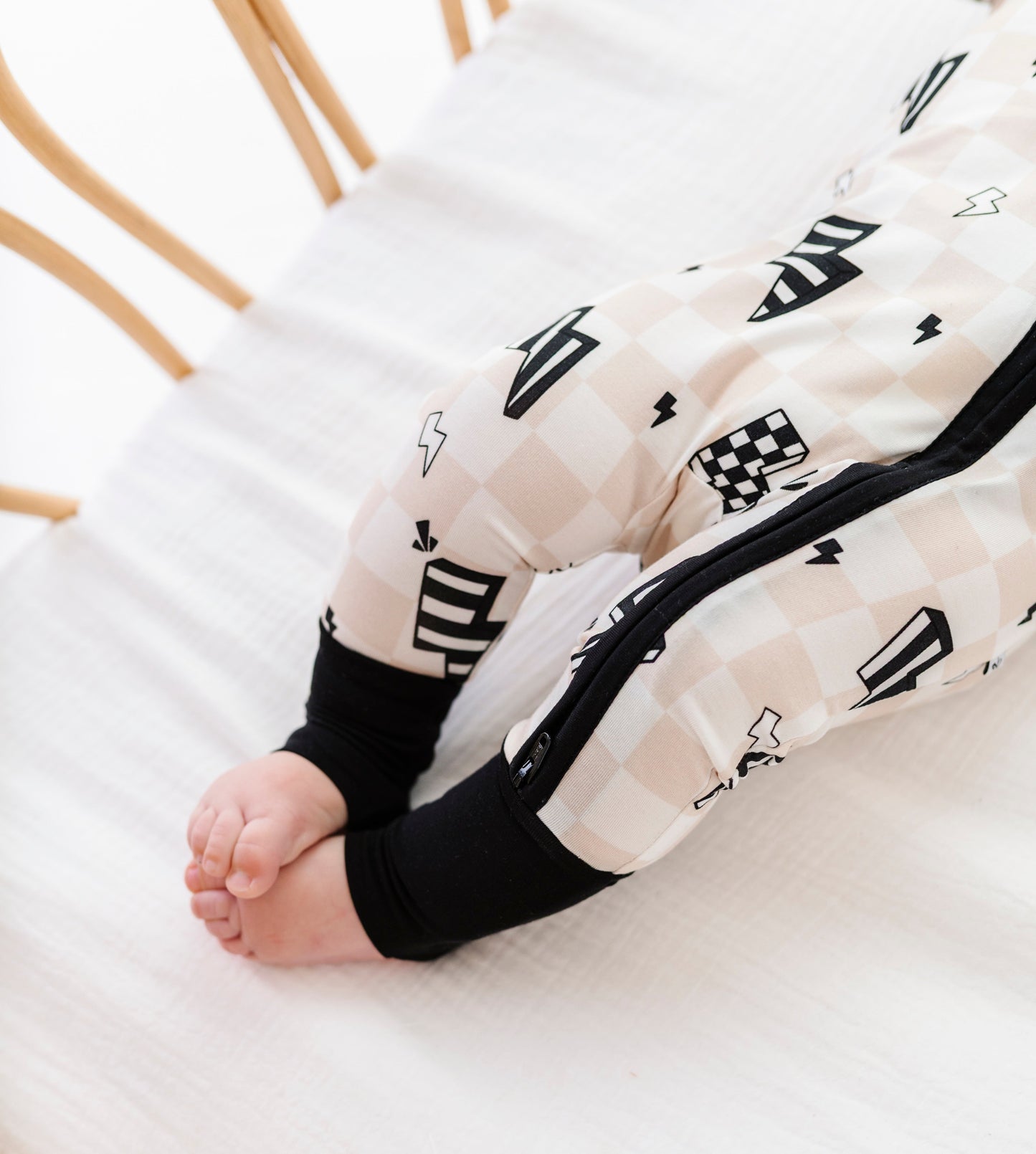 A baby's legs and feet wearing a cream and black checkered zip-up sleeper, lying on a white textured blanket inside a wooden crib.