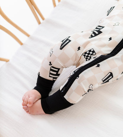 A baby's legs and feet wearing a cream and black checkered zip-up sleeper, lying on a white textured blanket inside a wooden crib.