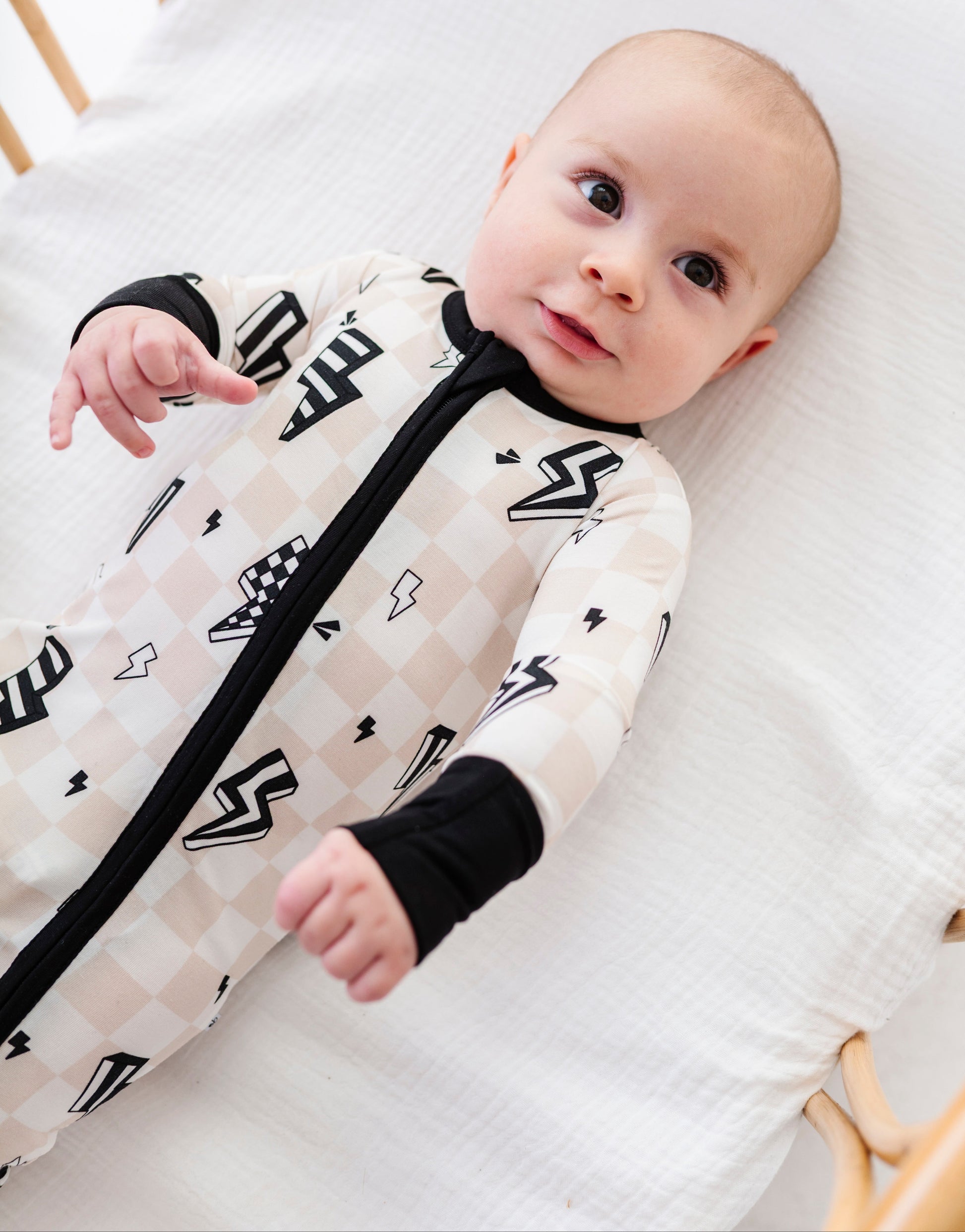 A baby lying on a white muslin mattress in a wooden bassinet, wearing a long-sleeve footless sleep sack with a black and white argyle pattern and black accents.