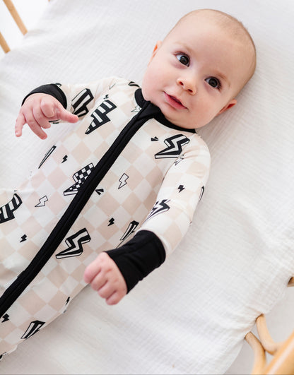 A baby lying on a white muslin mattress in a wooden bassinet, wearing a long-sleeve footless sleep sack with a black and white argyle pattern and black accents.