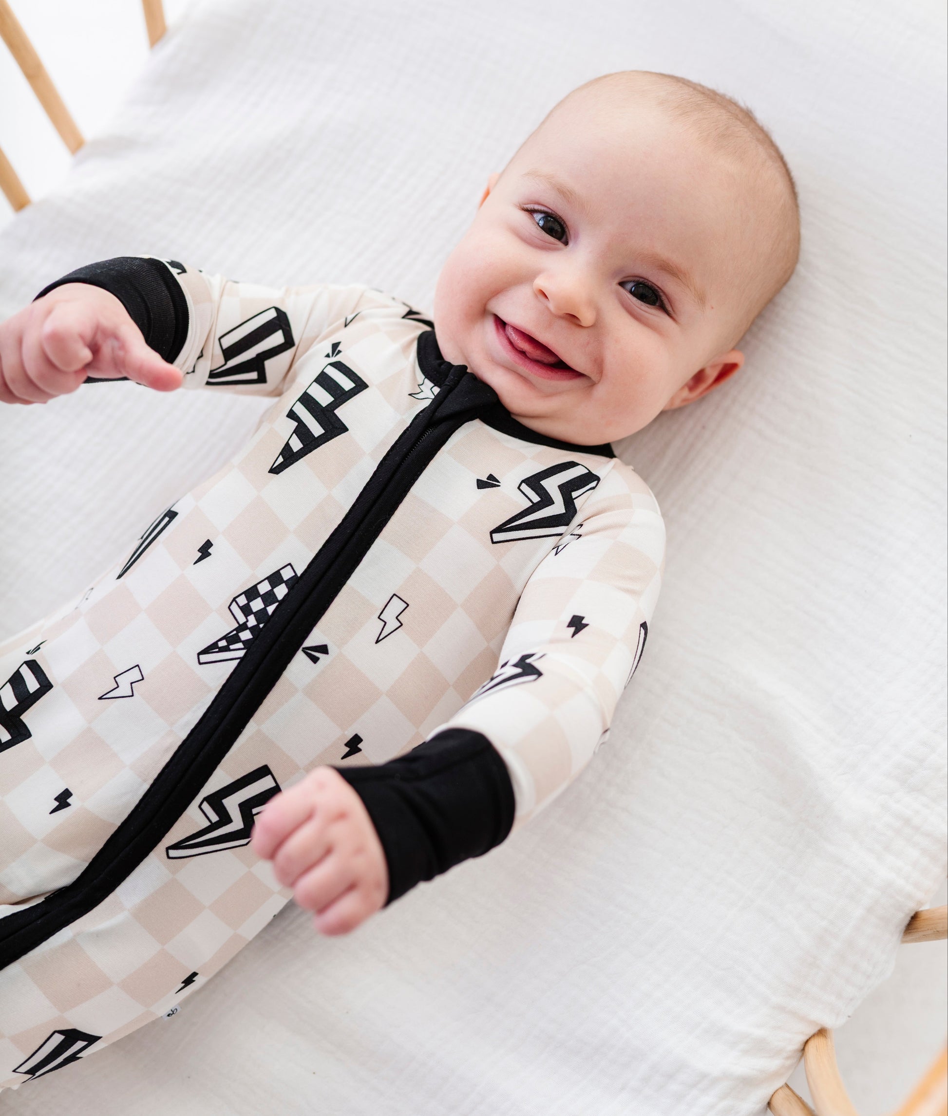 A baby lying on a white muslin mattress wearing a cream and beige checkered zip-up sleeper with black lightning bolt and checkerboard patterns, black cuffs, and a black zipper.