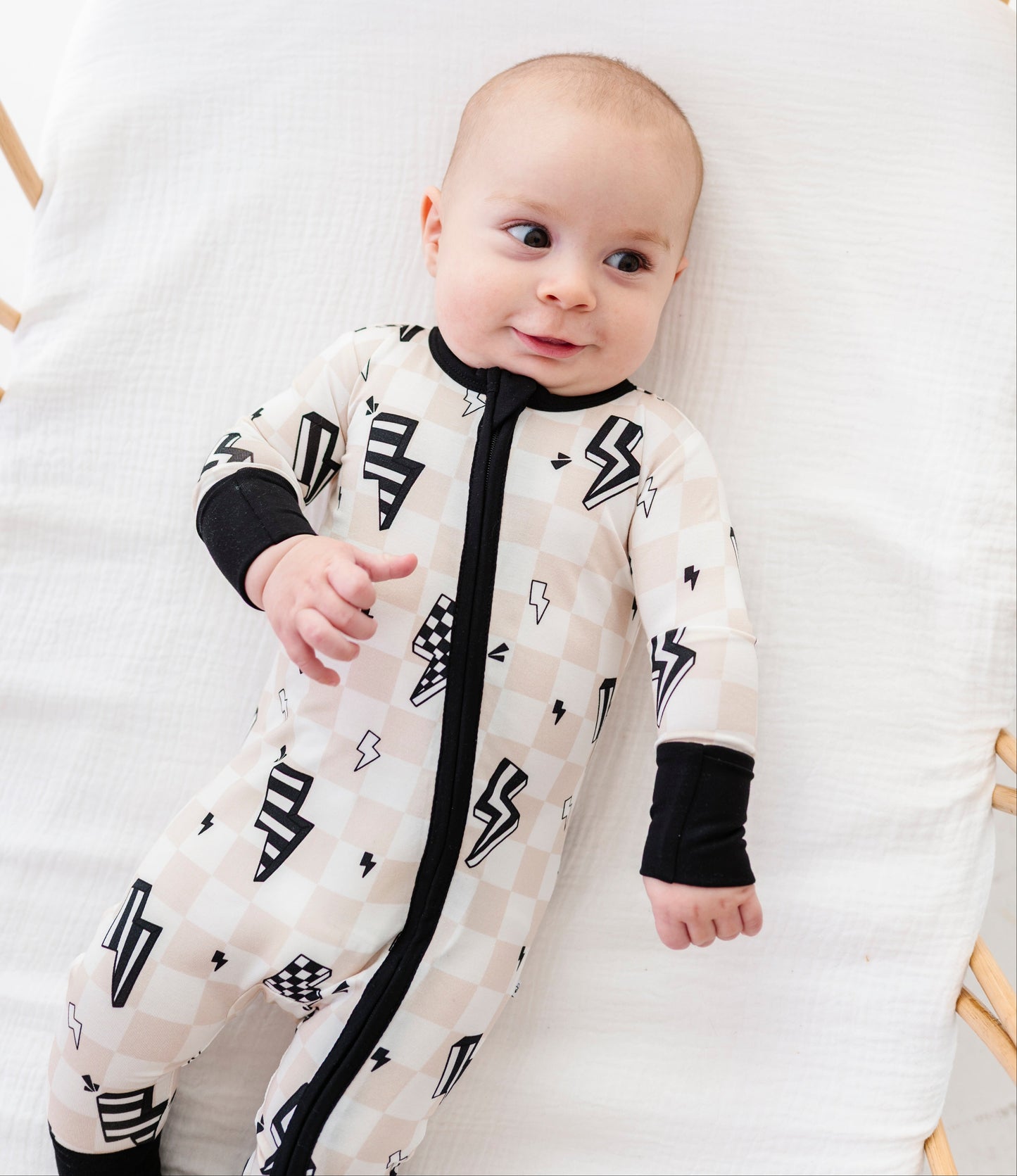 A baby lying on a white muslin blanket in a wooden bassinet, wearing a cream and beige checkered one-piece sleeper with black lightning bolt and checkerboard patterns, black cuffs, and a black front zipper.