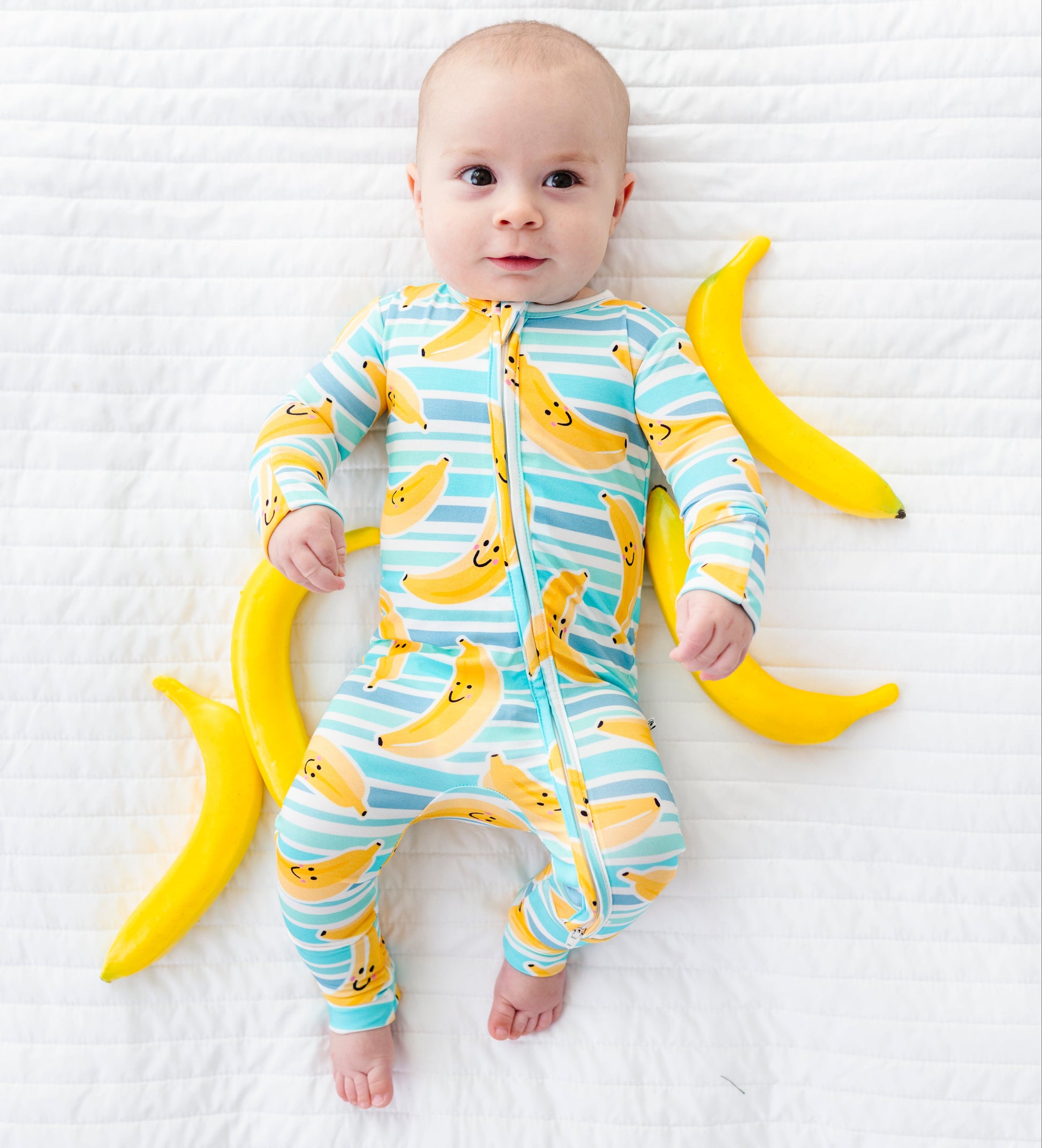 A baby lying on a white textured blanket, wearing a light blue and white striped one-piece pajama with a yellow banana print. Two yellow bananas are placed on either side of the baby as props.
