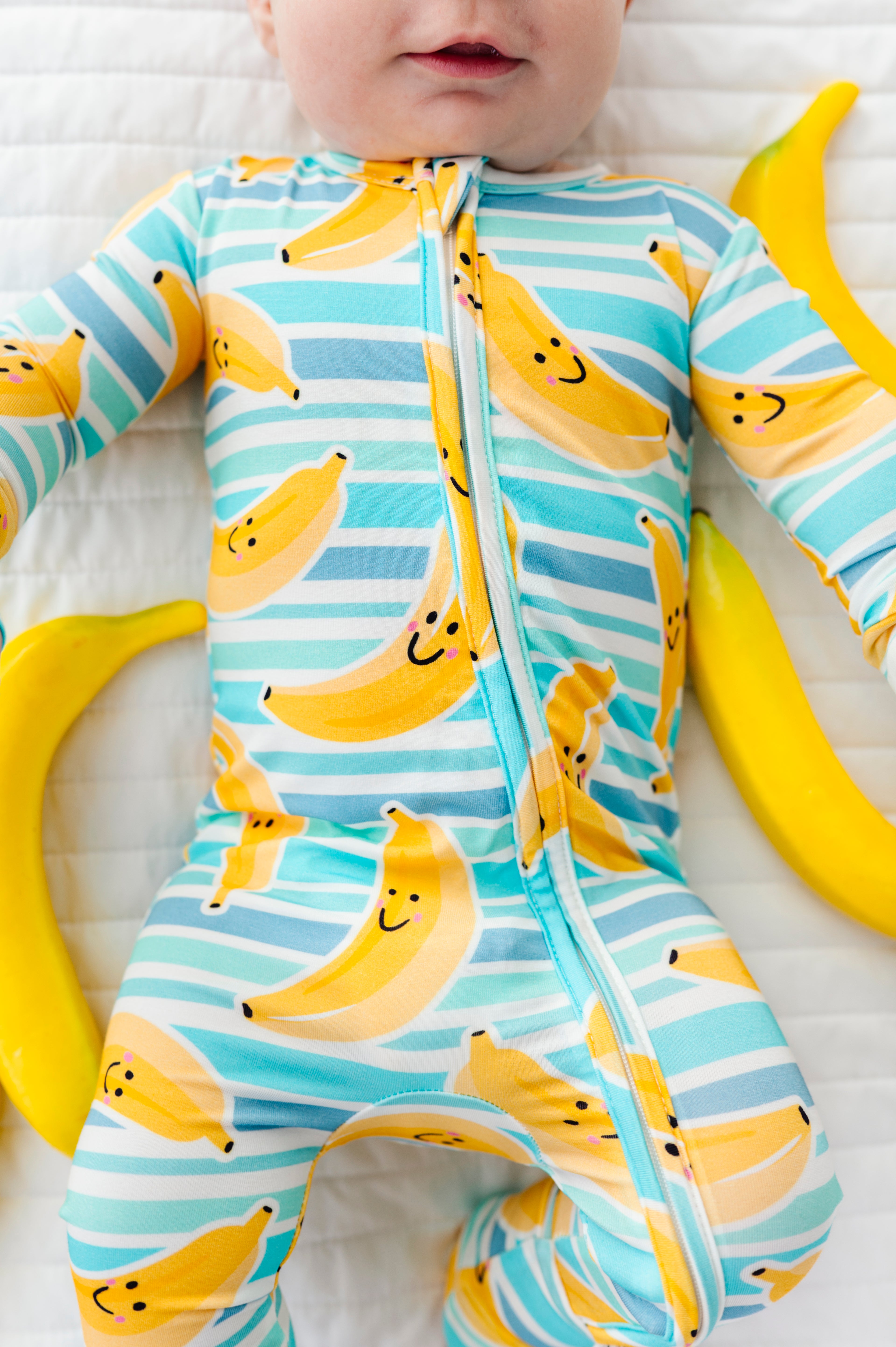 A baby lying on a white quilted blanket wearing a long-sleeve footless zip-up sleeper with a yellow banana print on a light blue and white striped background. Yellow banana props are placed around the baby.