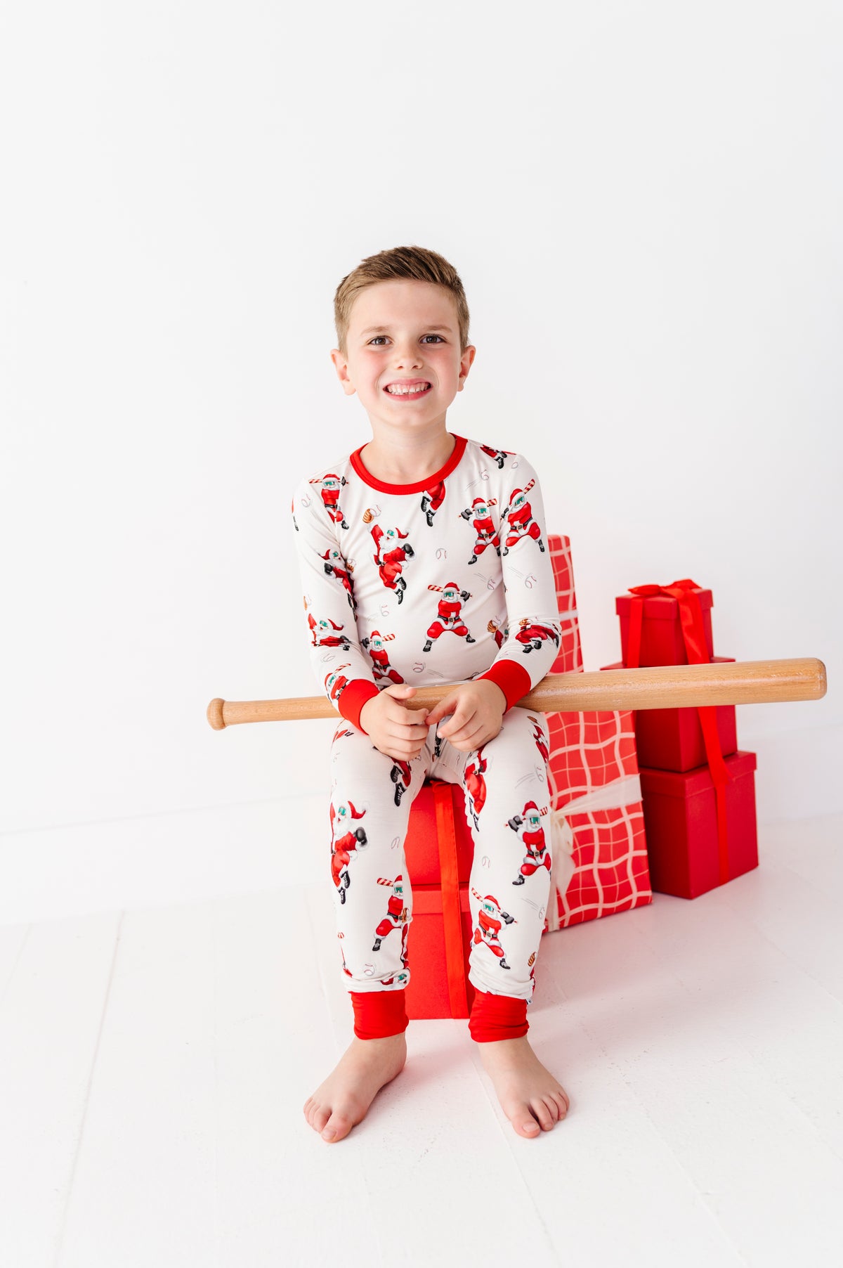 A young boy wearing white Christmas-themed pajamas with red Santa Claus prints sits on a red gift box, holding a wooden bat, with wrapped Christmas presents in the background against a white studio backdrop.