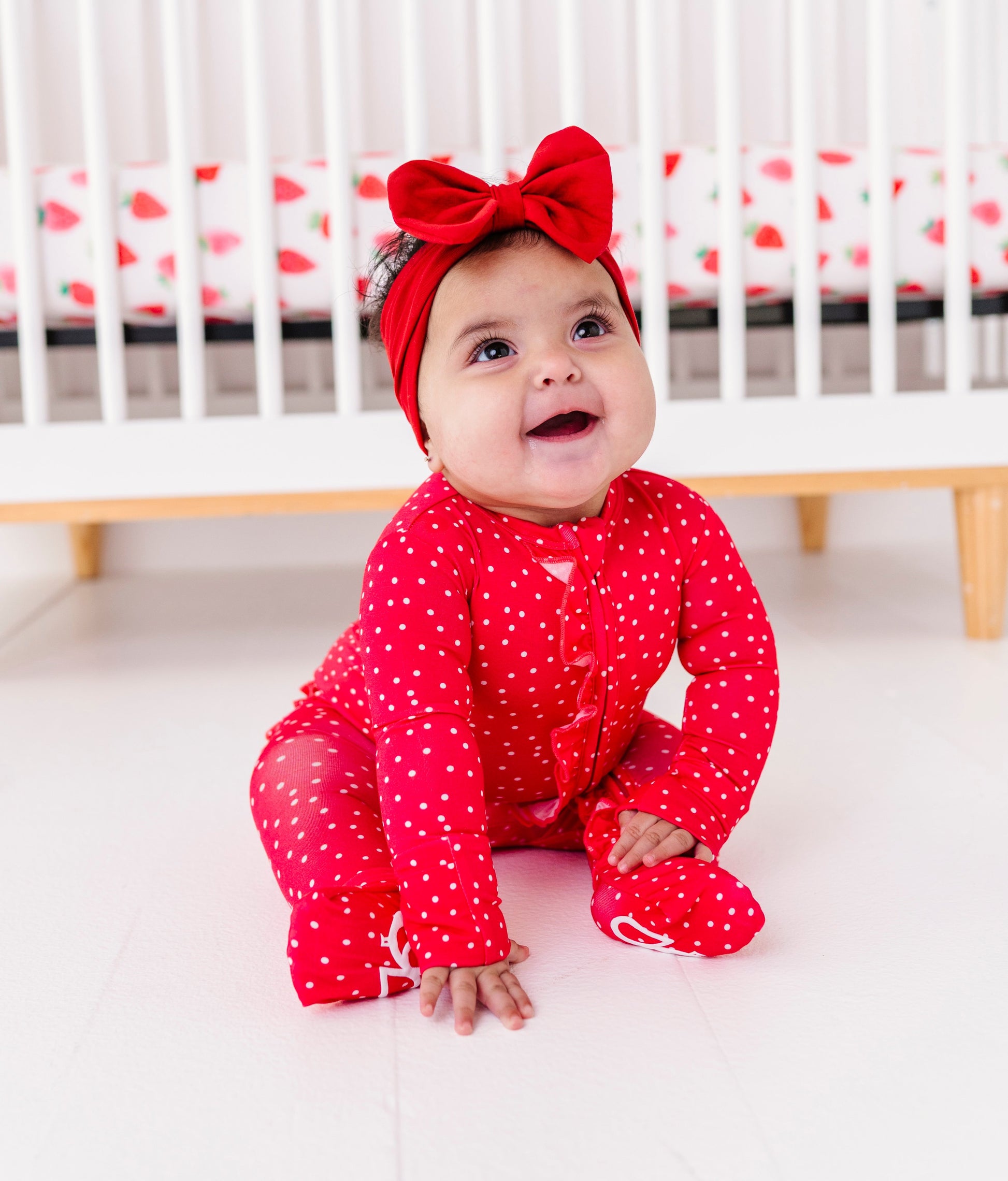 Baby in a red outfit with a bow sitting in front of a crib.
