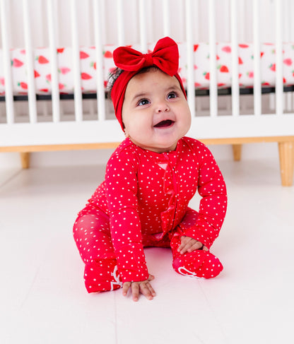 Baby in a red outfit with a bow sitting in front of a crib.