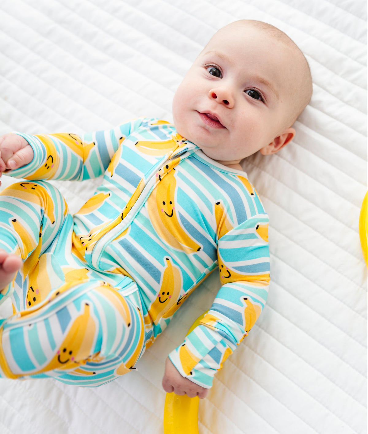A baby lying on a white quilted blanket wearing a long-sleeve zip-front onesie with a yellow banana print and light blue stripes.