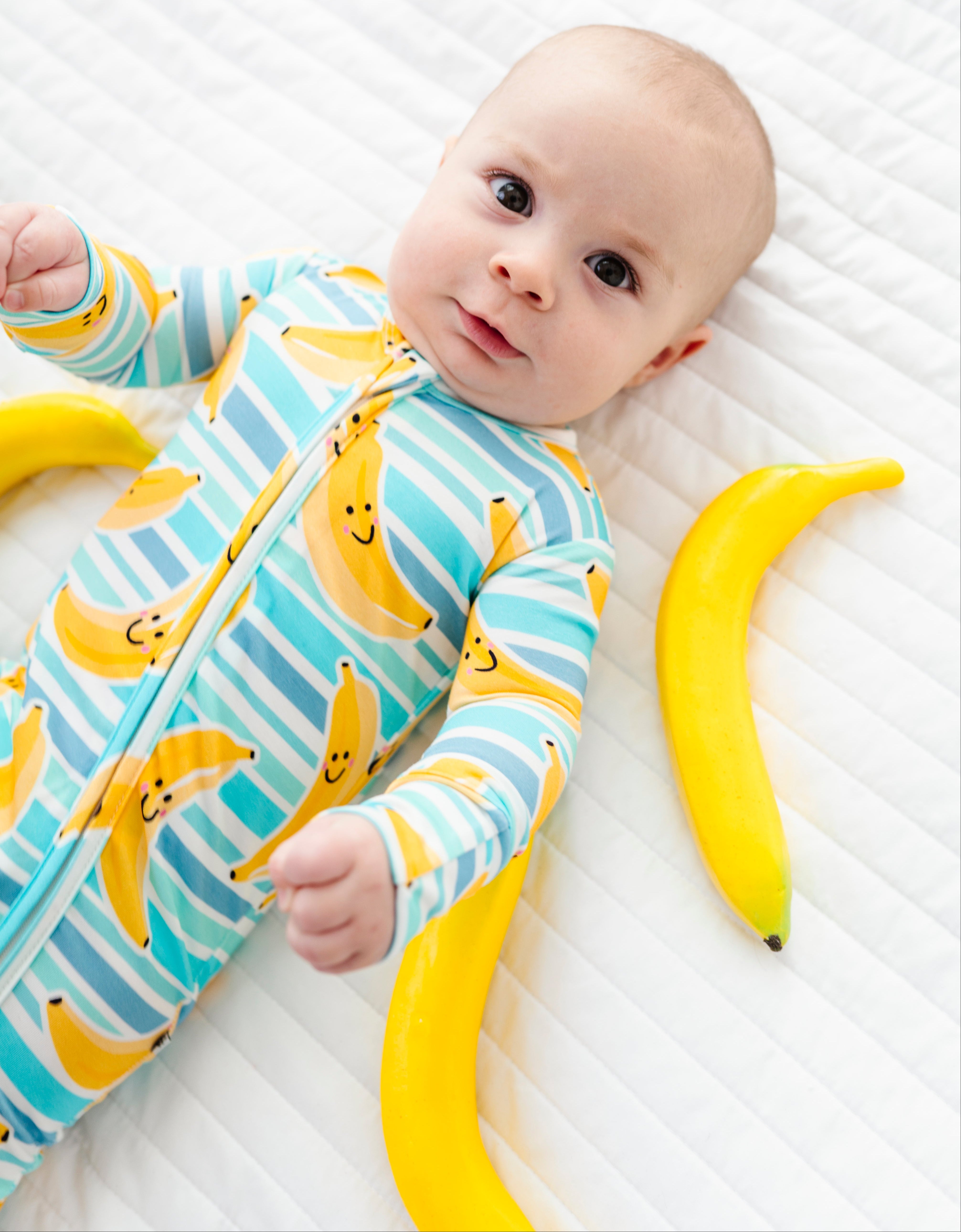 A baby lying on a white quilted mattress, wearing a light blue and white striped one-piece sleeper with a yellow banana print. Two large yellow bananas are placed on the mattress beside the baby.