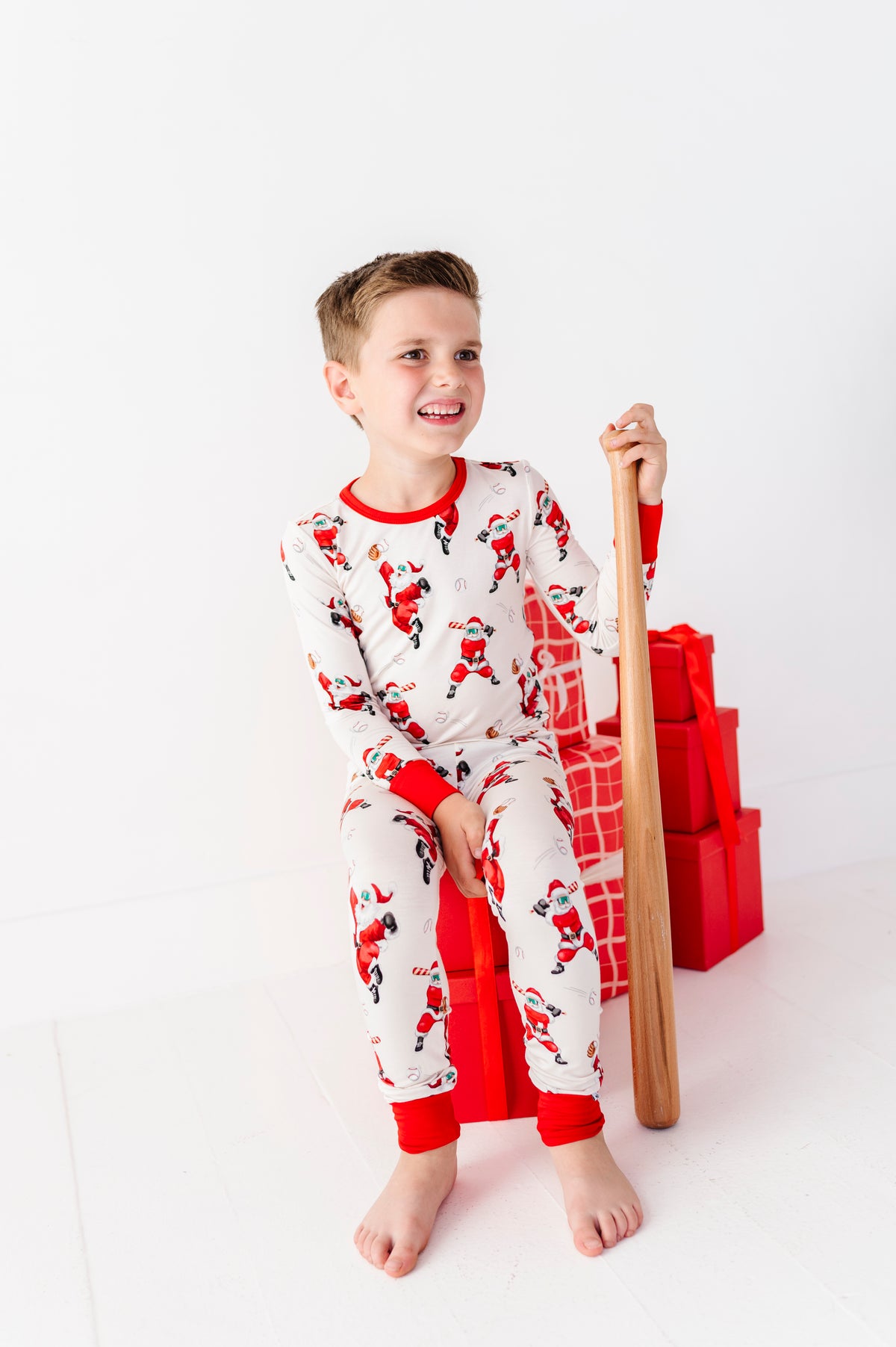 A young boy wearing bamboo pajamas featuring Santas playing baseball. He's sitting on a pile of red presents and holding a wooden baseball bat. 