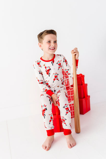 A young boy wearing bamboo pajamas featuring Santas playing baseball. He's sitting on a pile of red presents and holding a wooden baseball bat. 